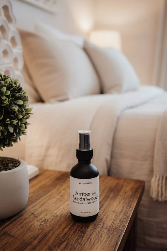 A bottle of KAT & GRACIE Amber and Sandalwood Room and Linen Spray, set on a wooden nightstand in a neutral bedroom