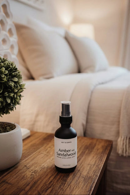 A bottle of KAT & GRACIE Amber and Sandalwood Room and Linen Spray, set on a wooden nightstand in a neutral bedroom