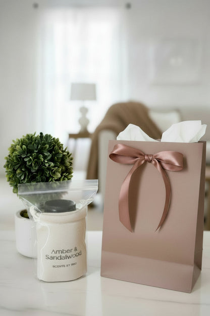 A packaged 'Amber & Sandalwood' scented sachet next to a gift bag and plant in a living room