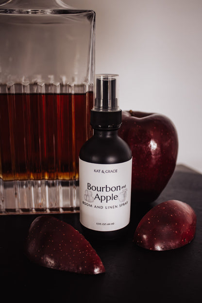 Bourbon and Apple room and linen spray bottle with apples and a glass of bourbon on a dark surface.
