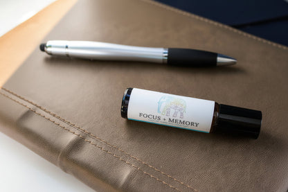 A rollerball bottle labeled 'FOCUS + MEMORY' with a white label and black cap, placed on a brown leather journal next to a silver and black pen
