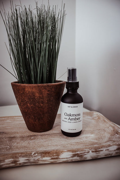 Bottle of 'Oakmoss & Amber' room spray next to a potted plant on a wooden surface.