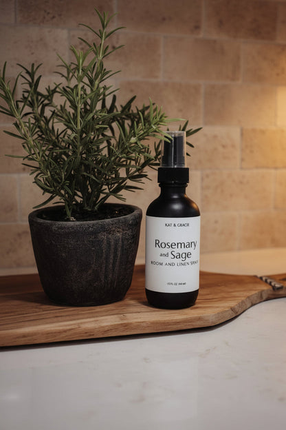 A bottle of KAT & GRACIE Rosemary and Sage Room and Linen Spray sits on a wooden cutting board on a marble countertop, next to a Rosemary plant with beige stone tile backsplash 