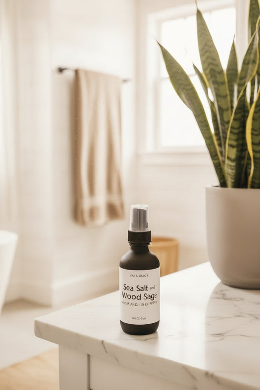 A bottle of Sea Salt and Wood Sage room and linen spray by Kat & Gracie, styled on a countertop next to a plant in a neutral bathroom
