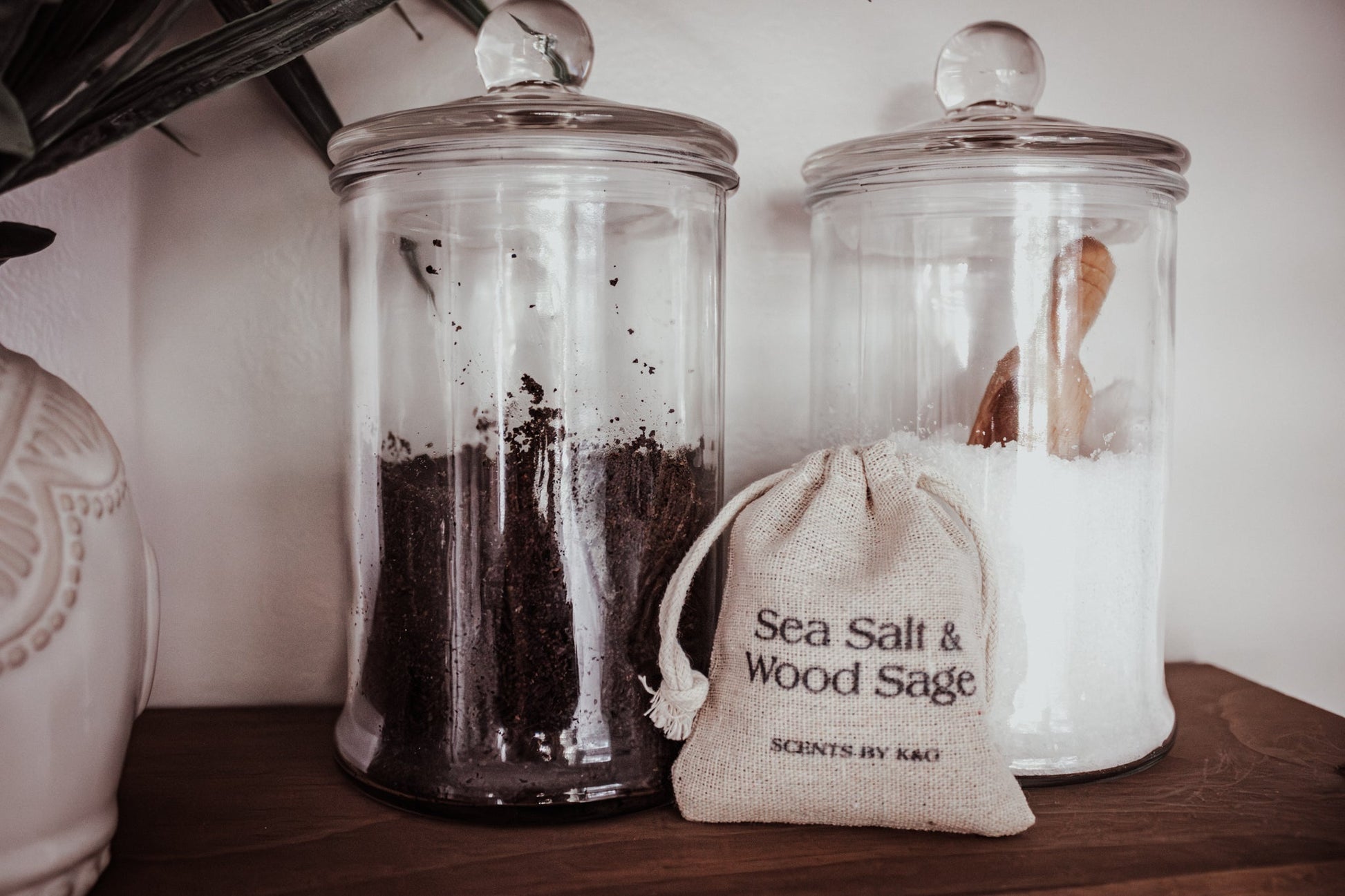 Two glass jars with lids and a small drawstring bag labeled 'Sea Salt & Wood Sage' on a wooden surface.