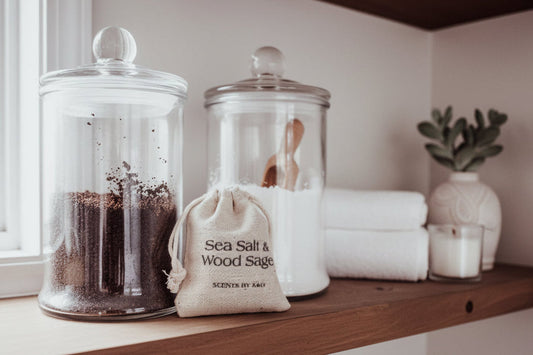 A 'Sea Salt & Wood Sage' scented sachet from Scents by K&G on a wood shelf with bath salts and coffee scrub next to towels and a plant