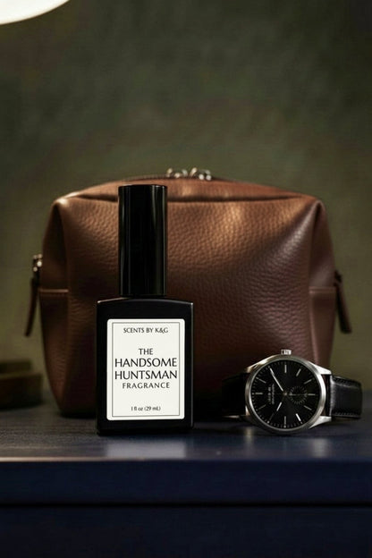 A bottle of Scents by K&G 'The Handsome Huntsman' fragrance stands in front of a brown leather zippered pouch, with a black-faced wristwatch resting beside it on a dark tabletop against a moody olive-green background.