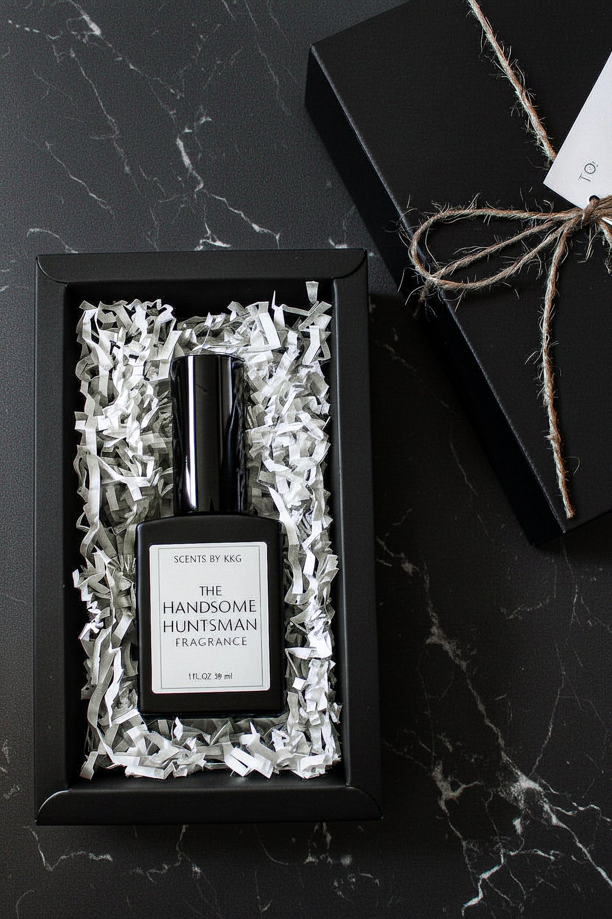 A black rectangular fragrance bottle labeled 'The Handsome Huntsman Fragrance' by Scents by K&G, presented in white crinkle paper inside a black gift box on a dark marble surface, with the box top and twine visible beside it.