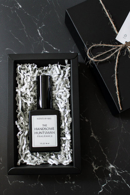 A black rectangular fragrance bottle labeled 'The Handsome Huntsman Fragrance' by Scents by K&G, presented in white crinkle paper inside a black gift box on a dark marble surface, with the box top and twine visible beside it.