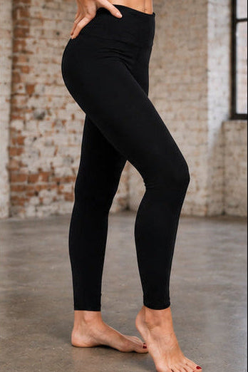 A person modeling black high-waisted leggings in a studio with a brick wall and concrete floor.