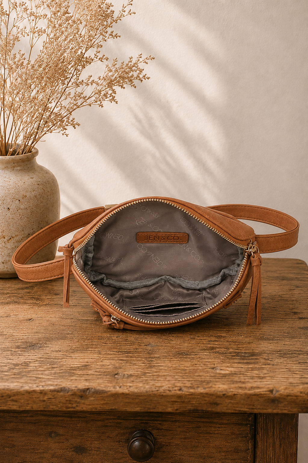 A tan leather crossbody bag with an open zipper, showing a gray interior lining, resting on a rustic wooden table next to a vase of dried flowers.
